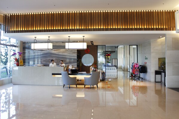 Una moderna zona de recepción con techo de listones de madera, mostrador de recepción, dos sillones grises y un amplio ventanal con luz natural.