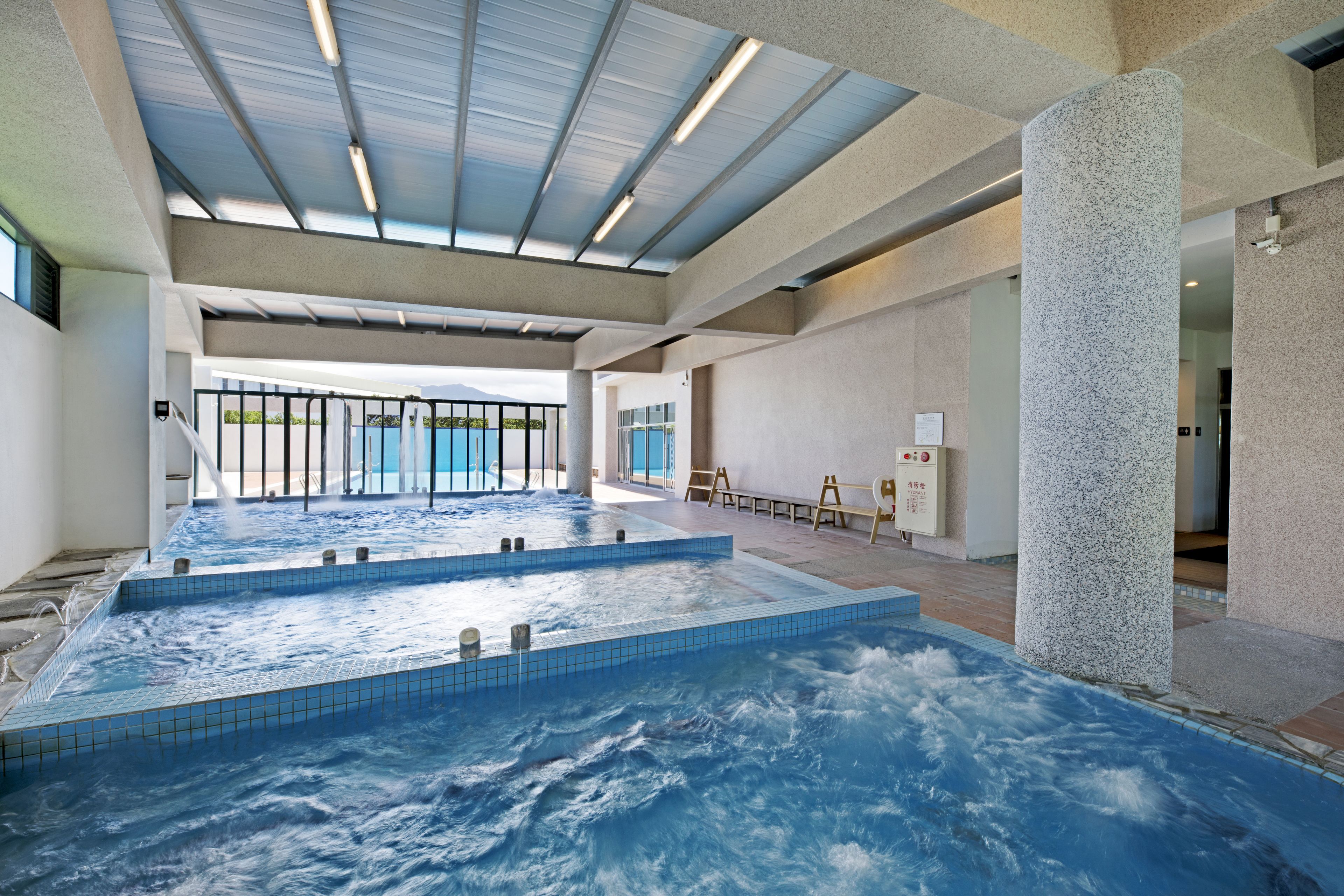 A large indoor swimming pool with multiple hot tubs, a skylight ceiling, and a view of the outdoors.