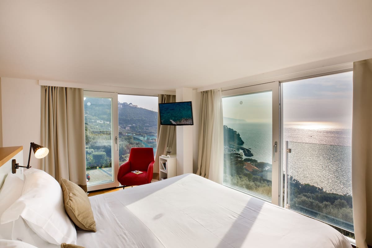 Camera d'hotel con un letto grande, una poltrona rossa, una TV e vista sul mare e sulle colline.