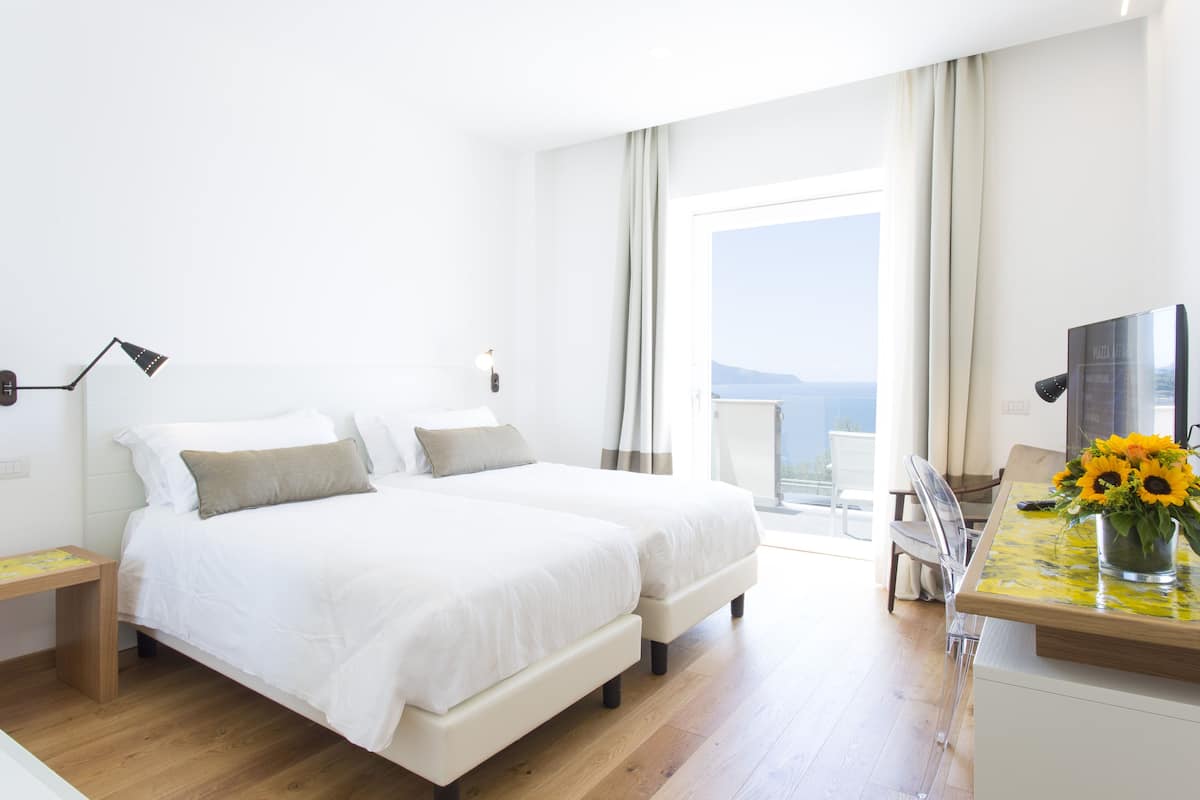 Una camera d'albergo con un letto, una scrivania con un vaso di girasoli e vista sul mare.