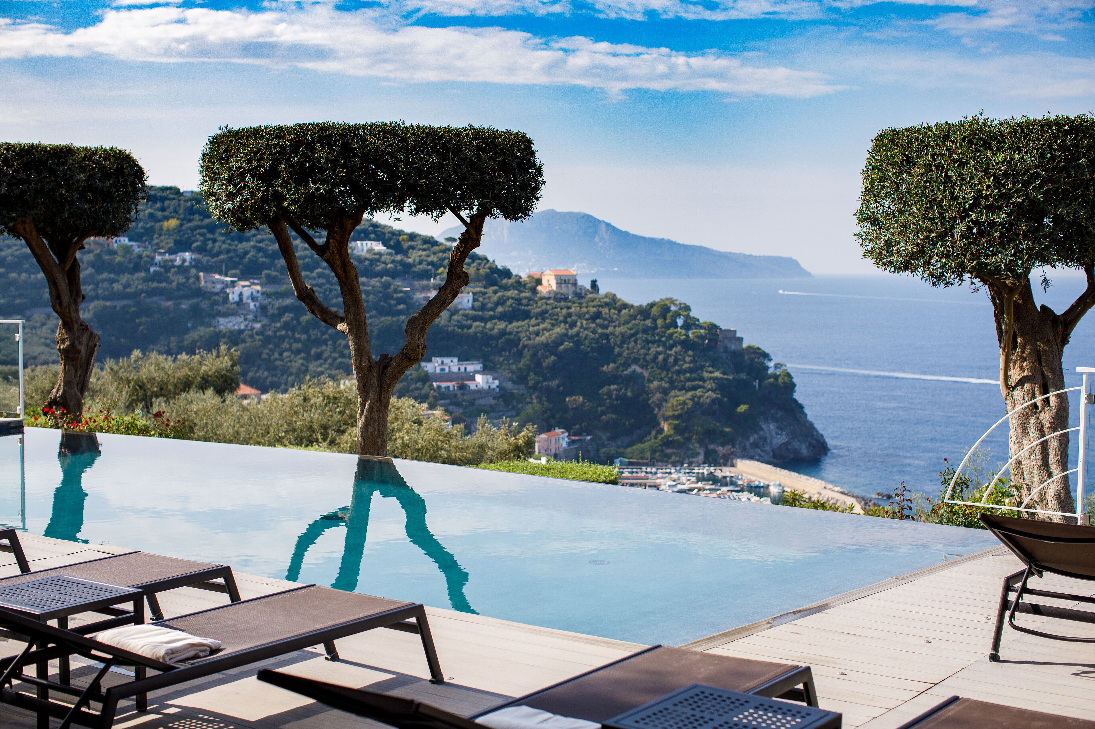 Une piscine à débordement avec des chaises longues, des arbres et une vue sur la mer et les bâtiments éloignés.
