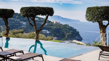 Une piscine à débordement avec des chaises longues, des arbres et une vue sur la mer et les bâtiments éloignés.