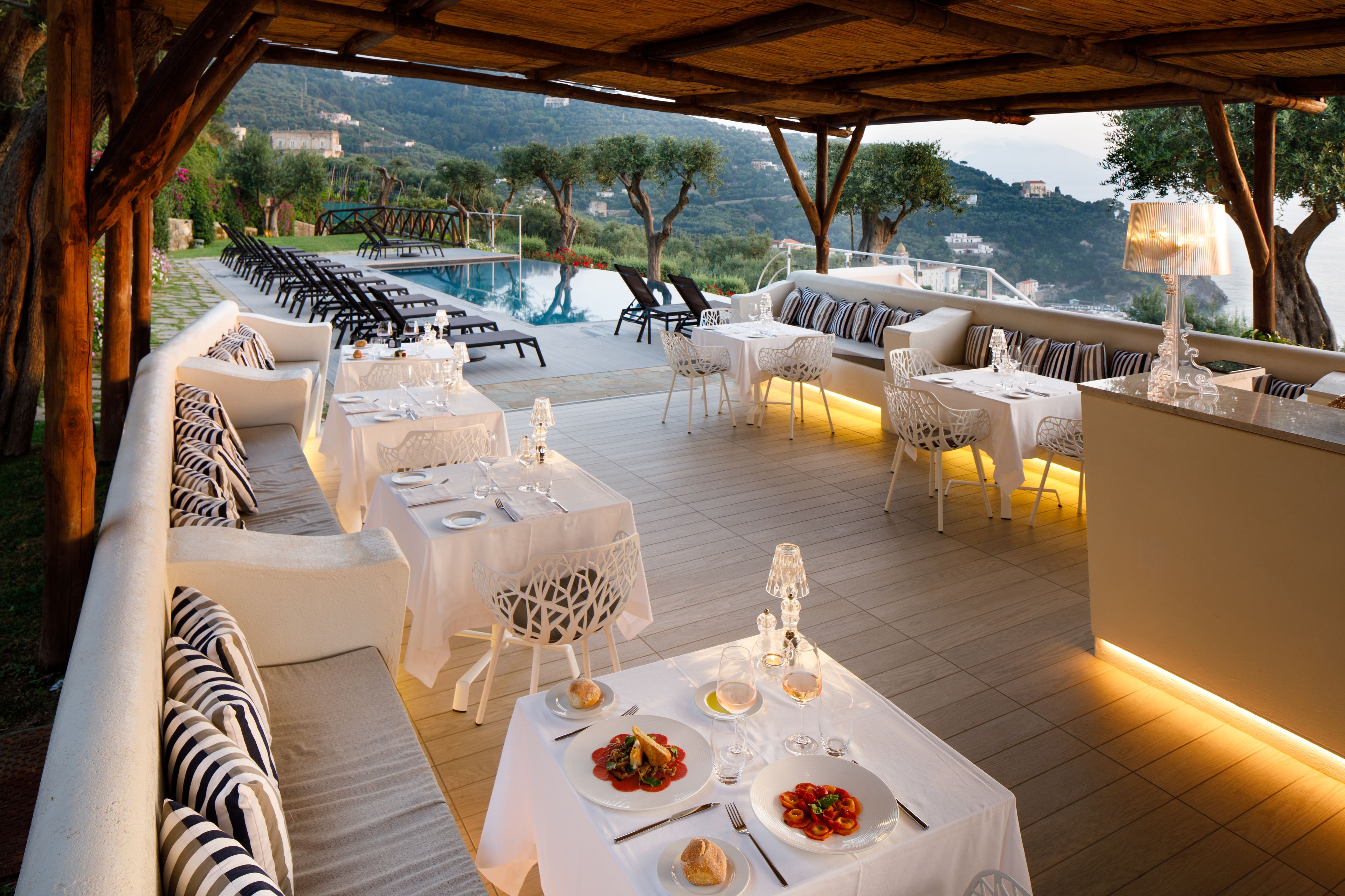 Un área de comedor al aire libre con mesas y sillas, una piscina y vistas del paisaje circundante.