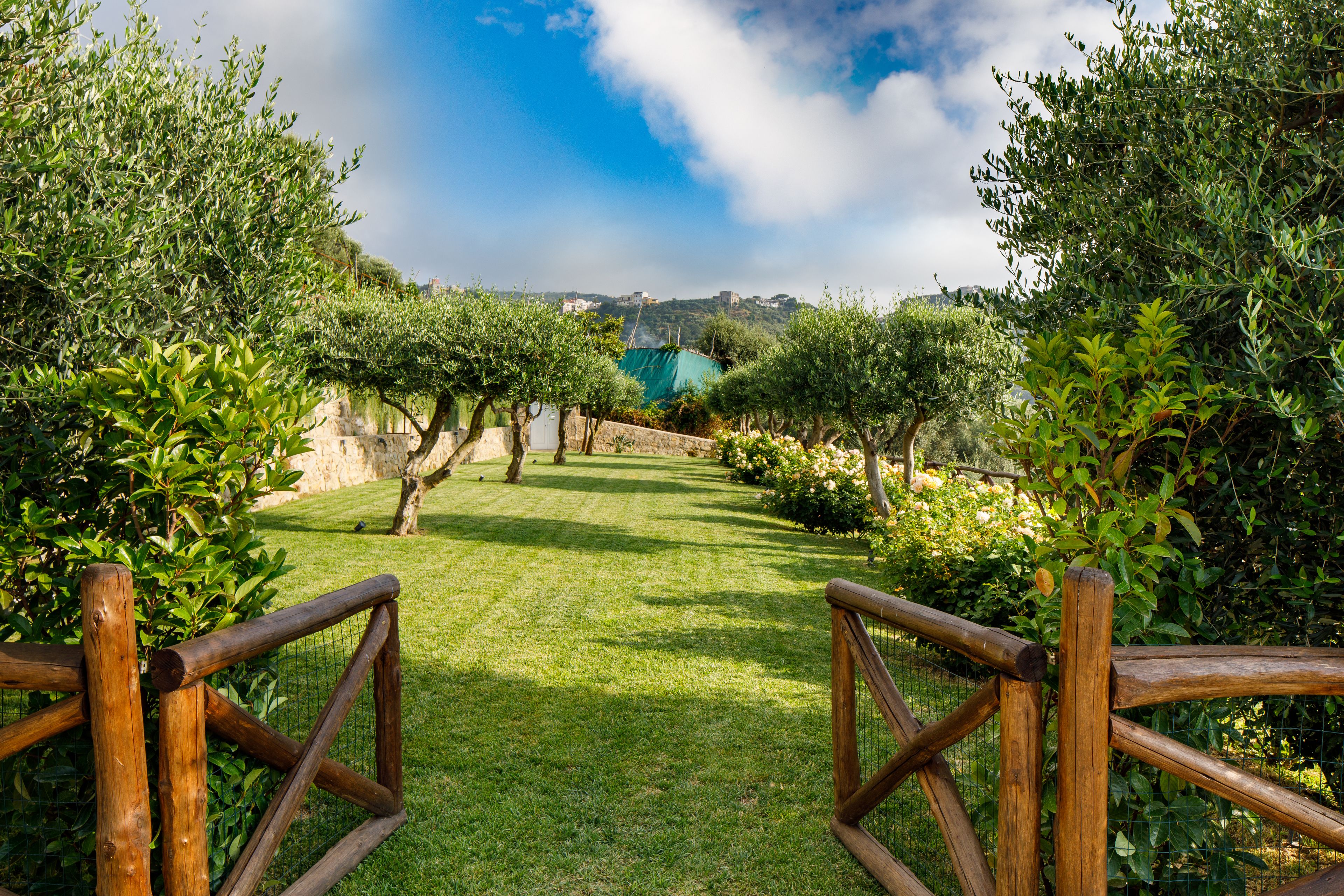 Un sentiero che si snoda in un giardino curato, con vista su una collina lontana e una costruzione blu.