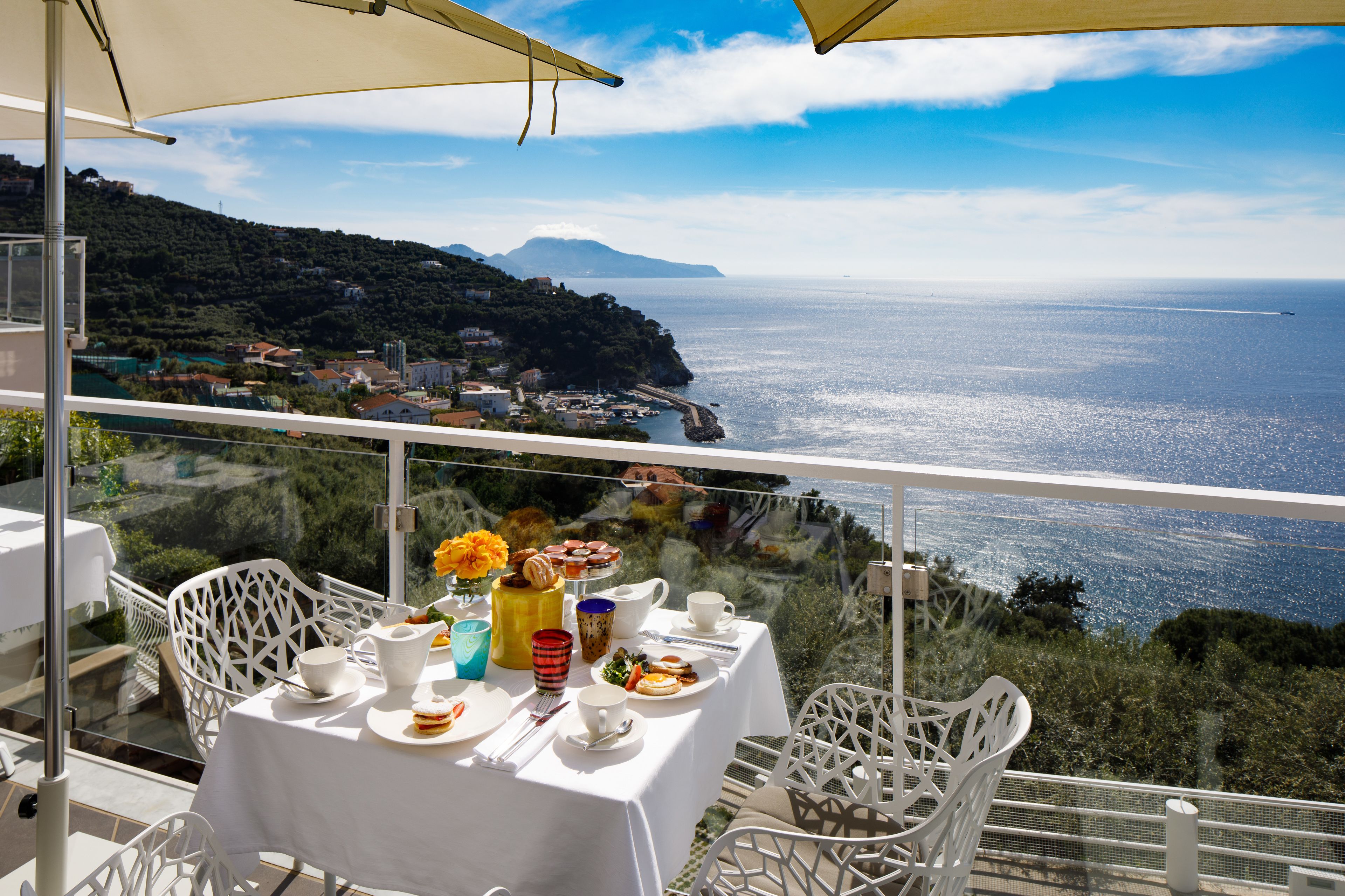 A table set for dining with a view of the sea and a mountainous landscape.