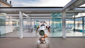 A modern outdoor dining area with glass walls, a wooden deck, and a view of the sea. - Villa Fiorella Art Hotel (Massa Lubrense)