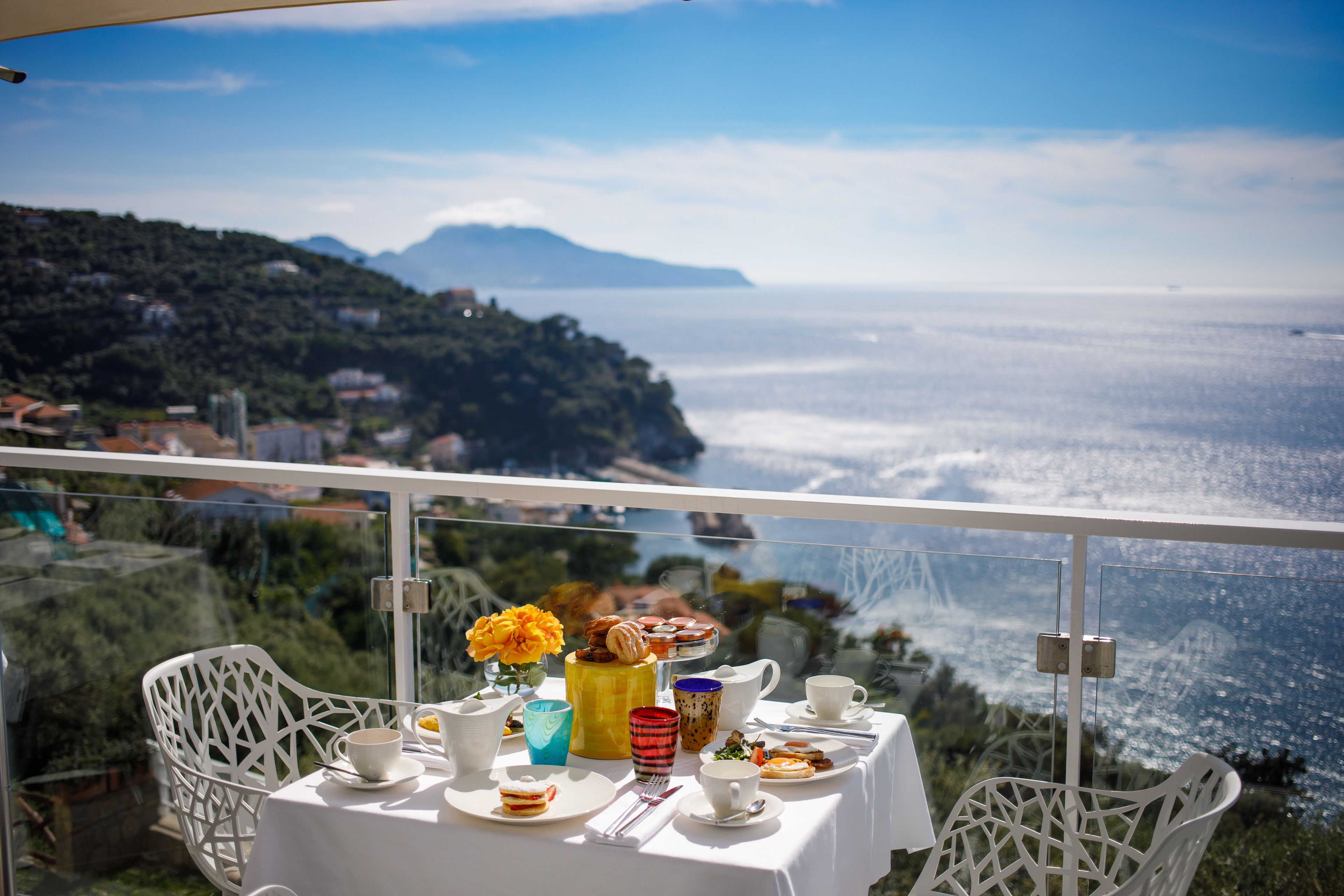 A table with a view of the sea and mountains, white chairs, and a table set with plates, cups, and a vase of flowers.