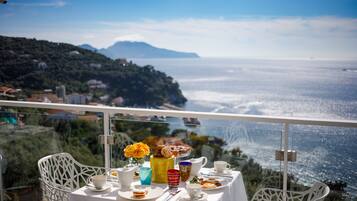 A table with a view of the sea and mountains, white chairs, and a table set with plates, cups, and a vase of flowers.
