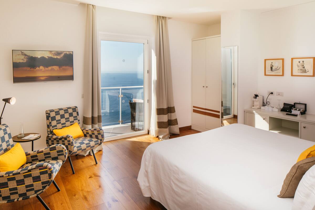 Camera d'albergo con un letto grande, due poltrone a scacchi con cuscini gialli, una scrivania con una sedia, una televisione a parete e un balcone con vista sul mare.