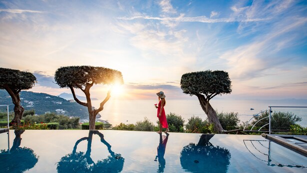 A person in a red dress standing by a reflective infinity pool with trees and a sunset in the background. - Villa Fiorella Art Hotel (Massa Lubrense)