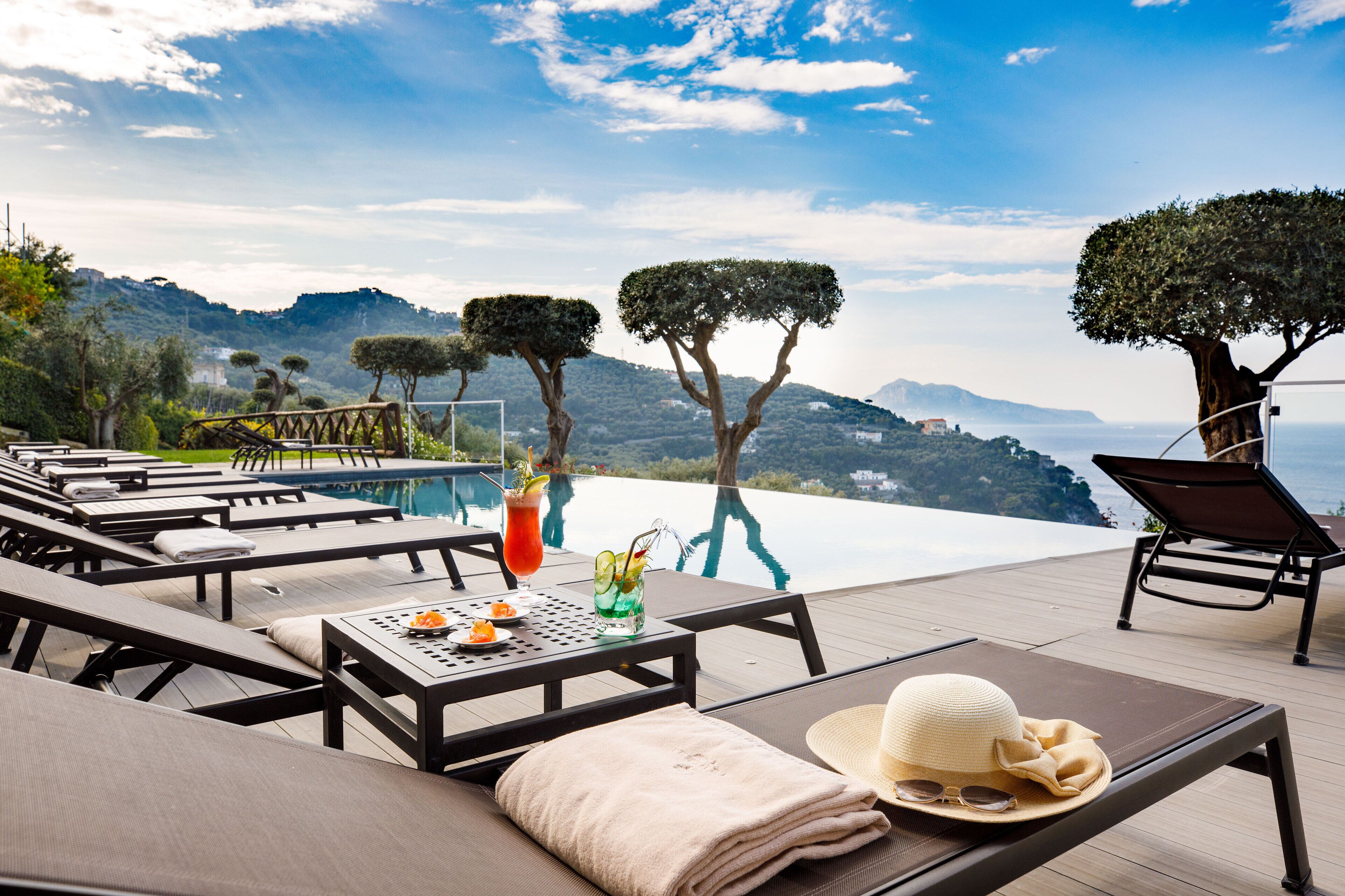 A poolside area with lounge chairs, a table with drinks, and a view of the sea and hills.