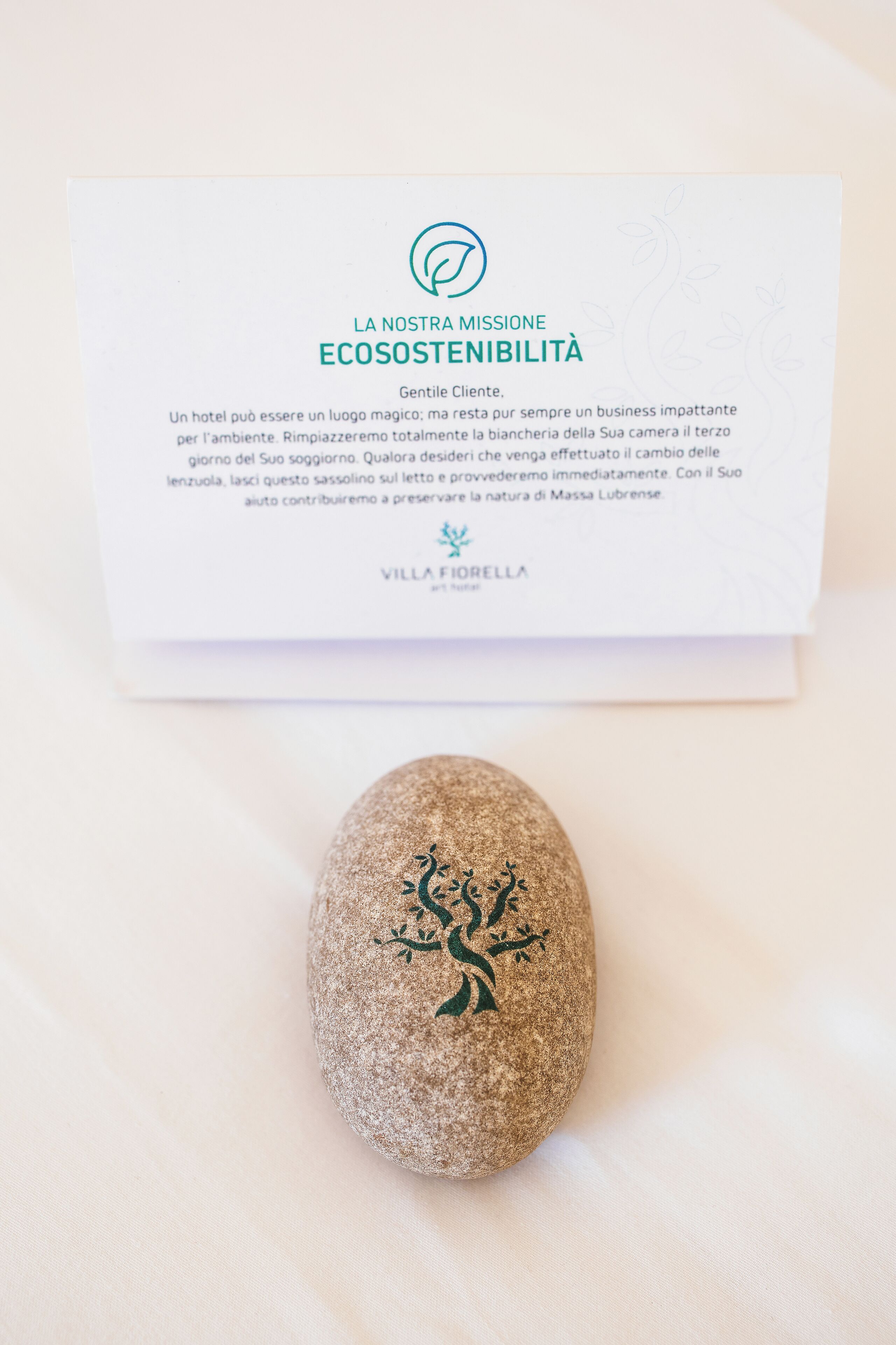 Een brochure met de titel 'LA NOSTRA MISSIONE ECOSOSTENIBILITÀ', een logo en tekst over milieuvriendelijke praktijken. Daaronder een steen met een ontwerp van een groene boom.