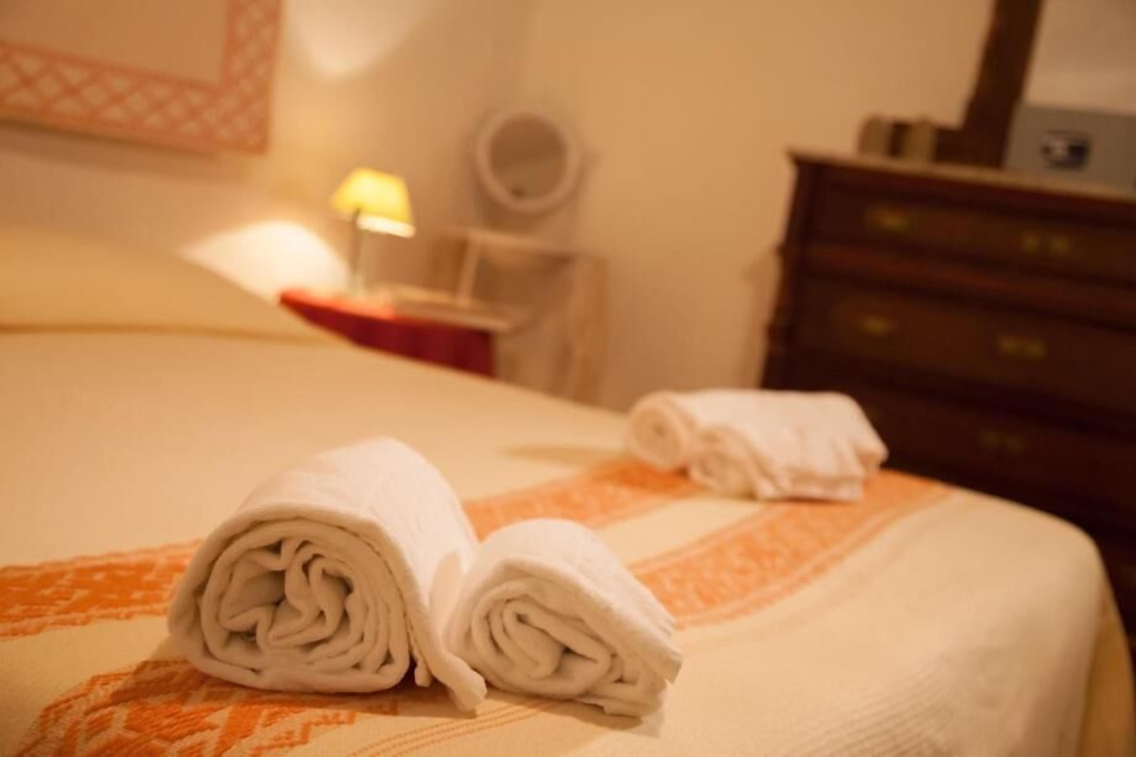 Una cama bien tendida con toallas blancas enrolladas y un aparador de madera al fondo.