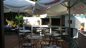 Un área de comedor al aire libre con mesas y sillas, cubierta con sombrillas, y una pared de ladrillo con una parrilla.
