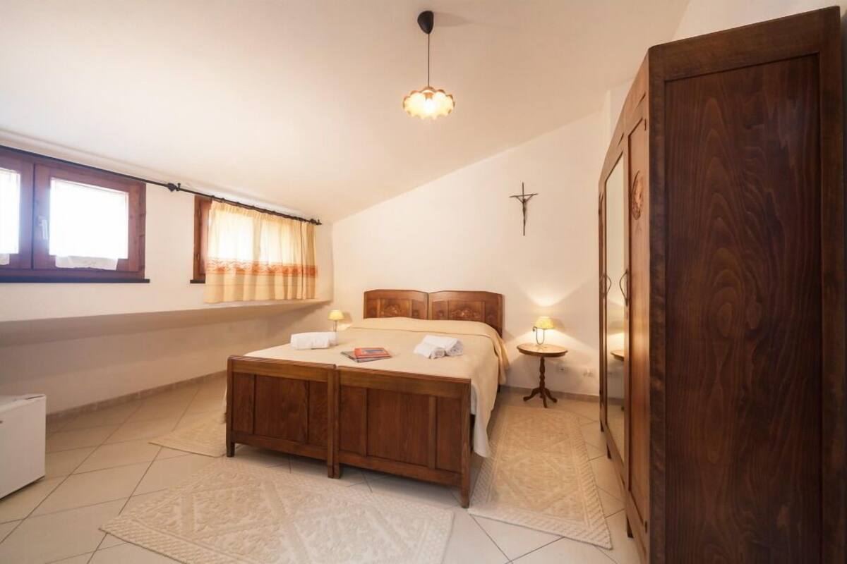 Un dormitorio con cama, mesitas de noche, un armario, una ventana con cortinas y una cruz en la pared.