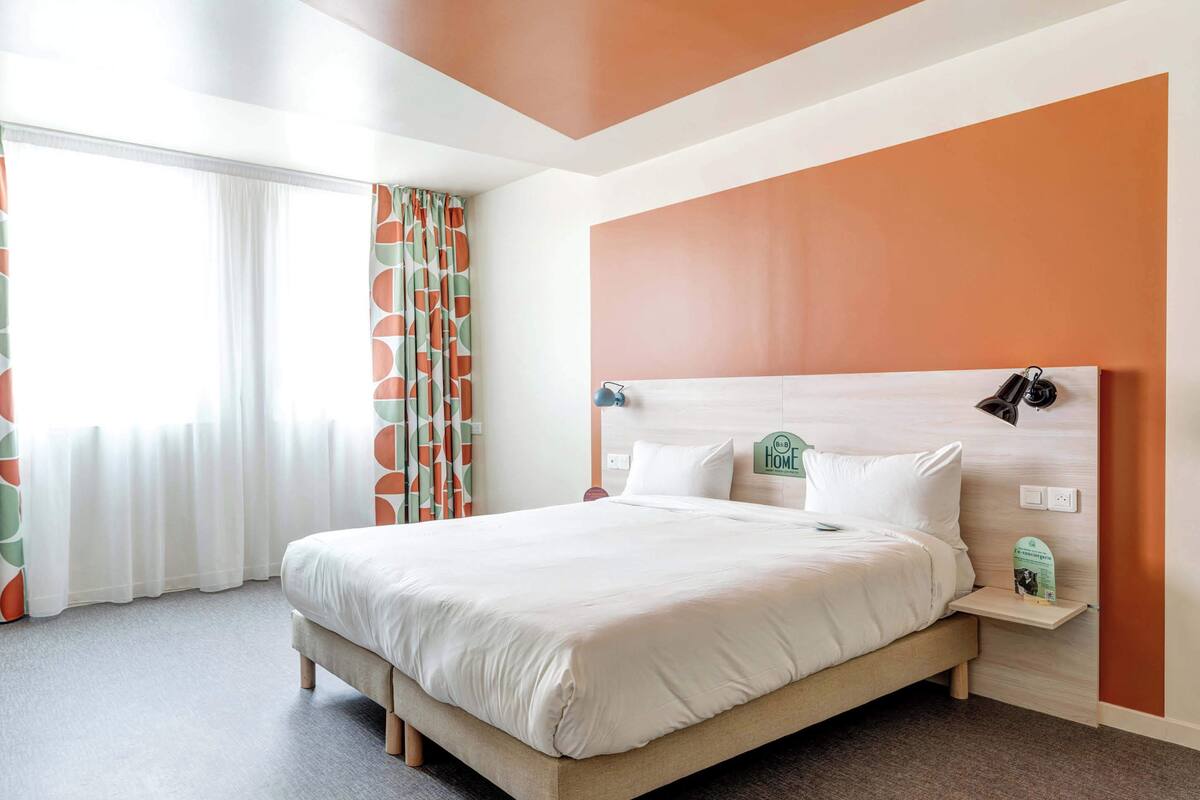 A neatly made bed in a room with a large window, a bedside table with a lamp, and a wall-mounted clock.