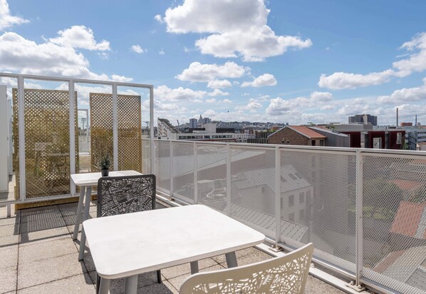 A rooftop terrace with a table and chairs, a view of the cityscape, and a mesh fence.