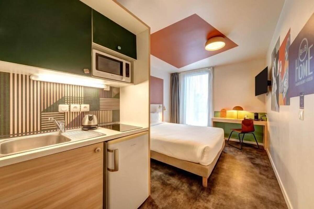 A compact hotel room with a kitchenette, a bed, a desk, and a chair.
