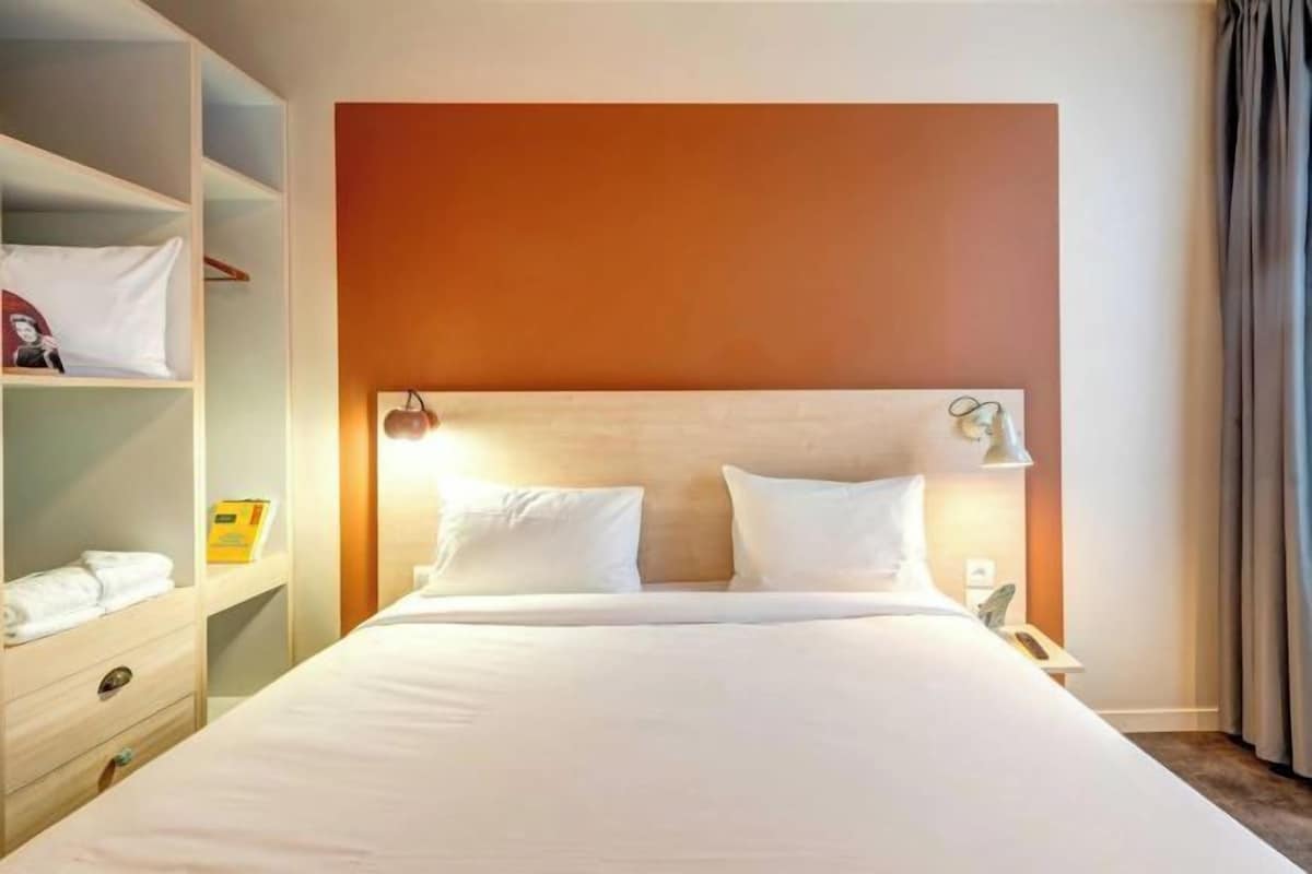 Ein Hotelzimmer mit einem großen Bett, einem Holzkopfteil, einem Nachttisch mit Lampe und einem Kleiderschrank mit darin hängender Kleidung.