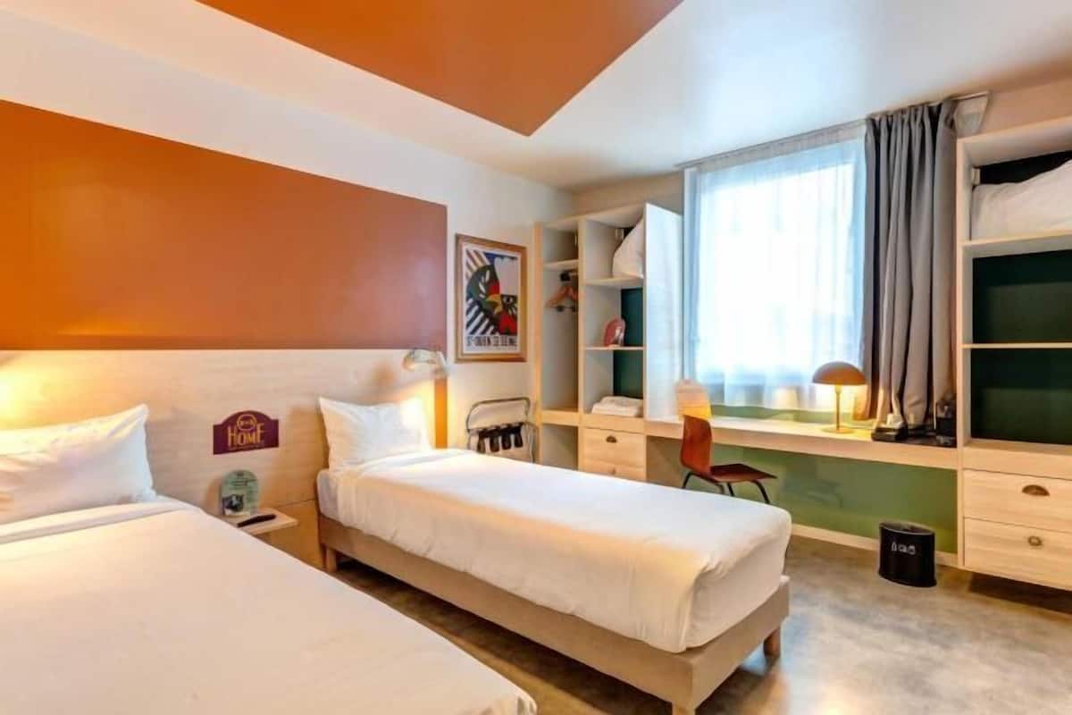A hotel room with two beds, a desk, a chair, a window with curtains, and a trash bin.