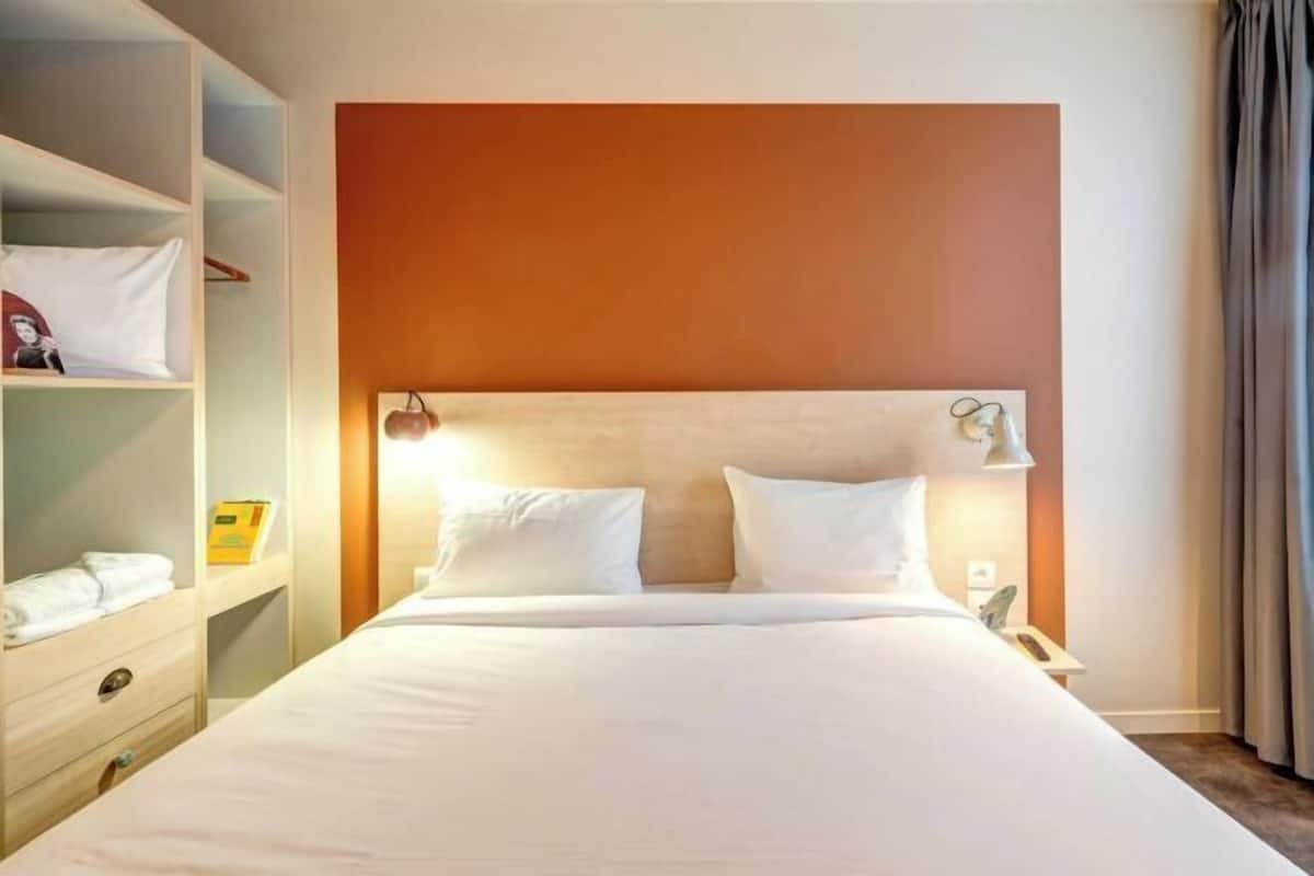 A hotel room with a large bed, a wooden headboard, a bedside table with a lamp, and a wardrobe with hanging clothes.