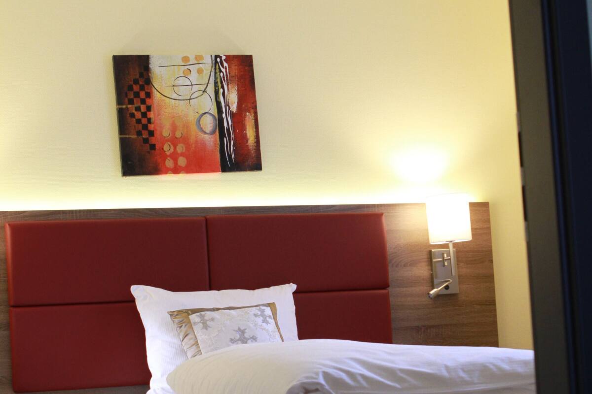Habitación de hotel con cama, cabecero rojo, lámpara de pared y una obra de arte abstracta.