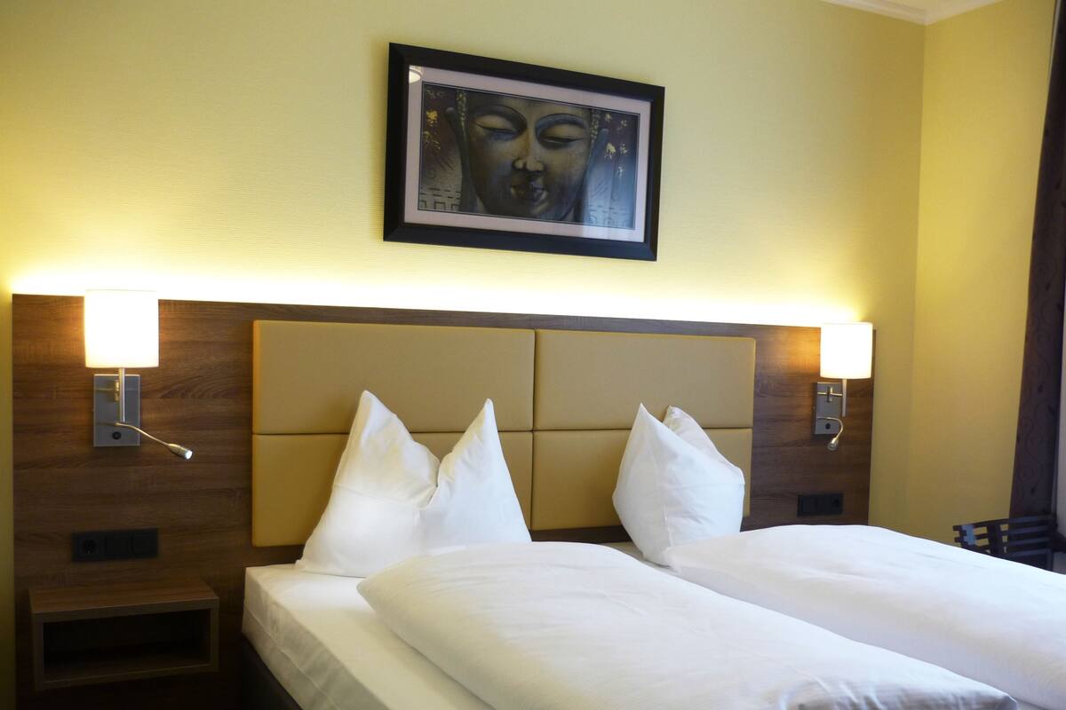 Una habitación de hotel con una cama grande, dos lámparas de noche, una obra de arte enmarcada sobre la cama y una mesita de noche de madera.