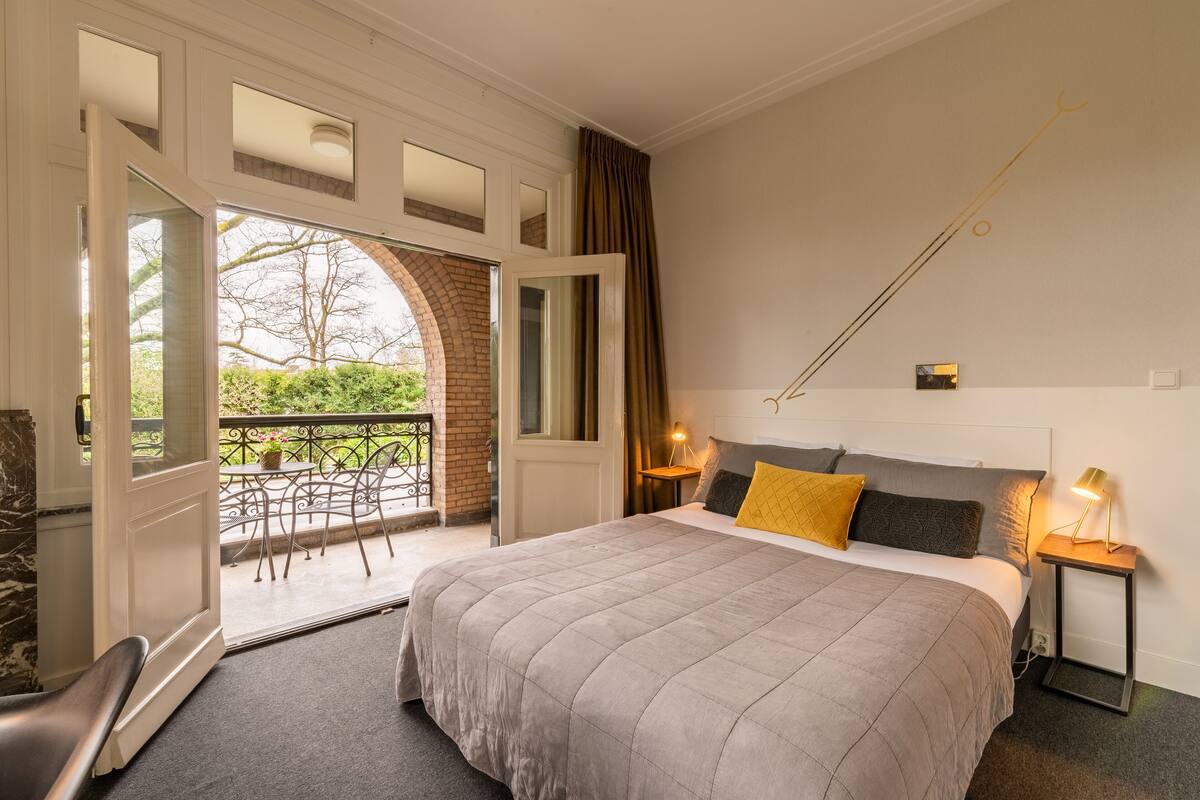 Une chambre avec un grand lit, un balcon, une vue sur la verdure et une petite table avec une lampe.
