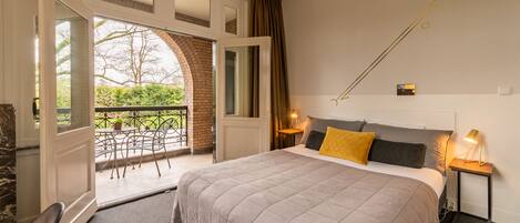 A bedroom with a large bed, a balcony, a view of greenery, and a small table with a lamp.