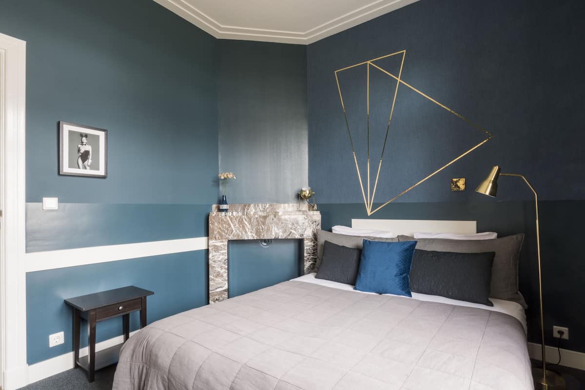 Une chambre dotée d’un grand lit, d’une table de chevet, d’une cheminée en marbre et d’un luminaire moderne aux formes géométriques.