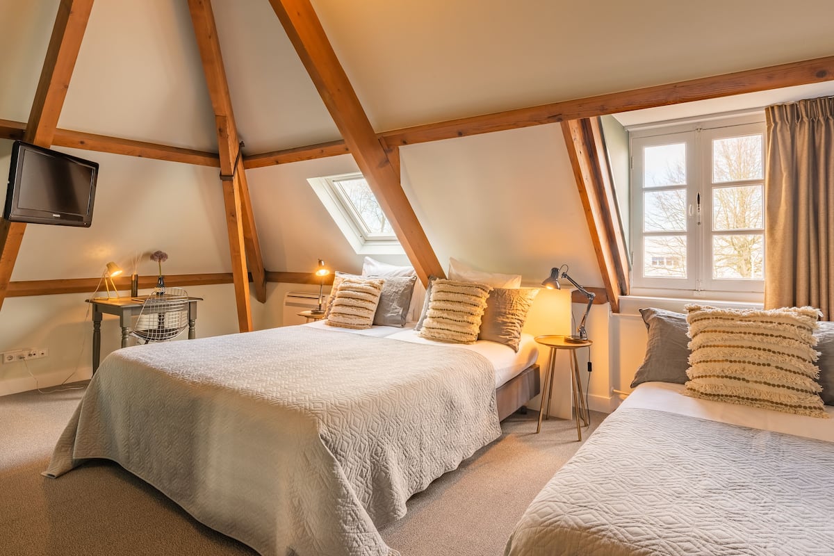 Une chambre avec deux lits, des poutres en bois, une télévision, une fenêtre avec des rideaux et une lampe.