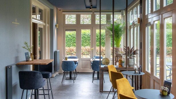 A modern interior with a dining area featuring blue and yellow chairs, a wooden table, and large windows offering a view of greenery outside. - Boutique Hotel Het Scheepshuys (Breda)