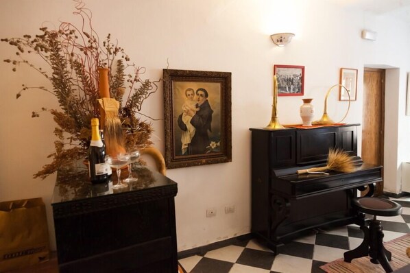 A room with a checkered floor, a black piano, a framed picture, and a vase with dried plants.