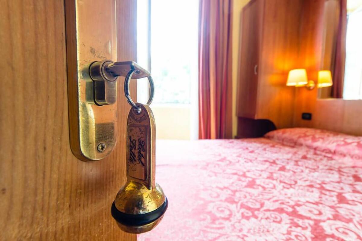 Quarto de hotel com porta de madeira, chaveiro, cama com colcha estampada e luminária fixada na parede.