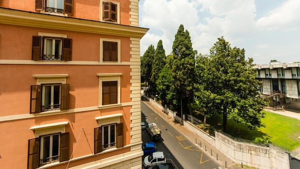 A multi-story building with balconies, a road with vehicles, and a green area with trees. - Hotel Lella (Rome)