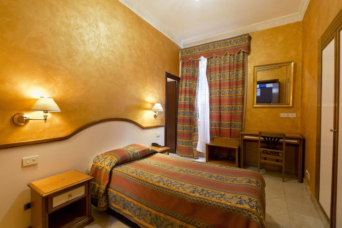 Quarto de hotel com cama, luminárias de cabeceira, escrivaninha e espelho.
