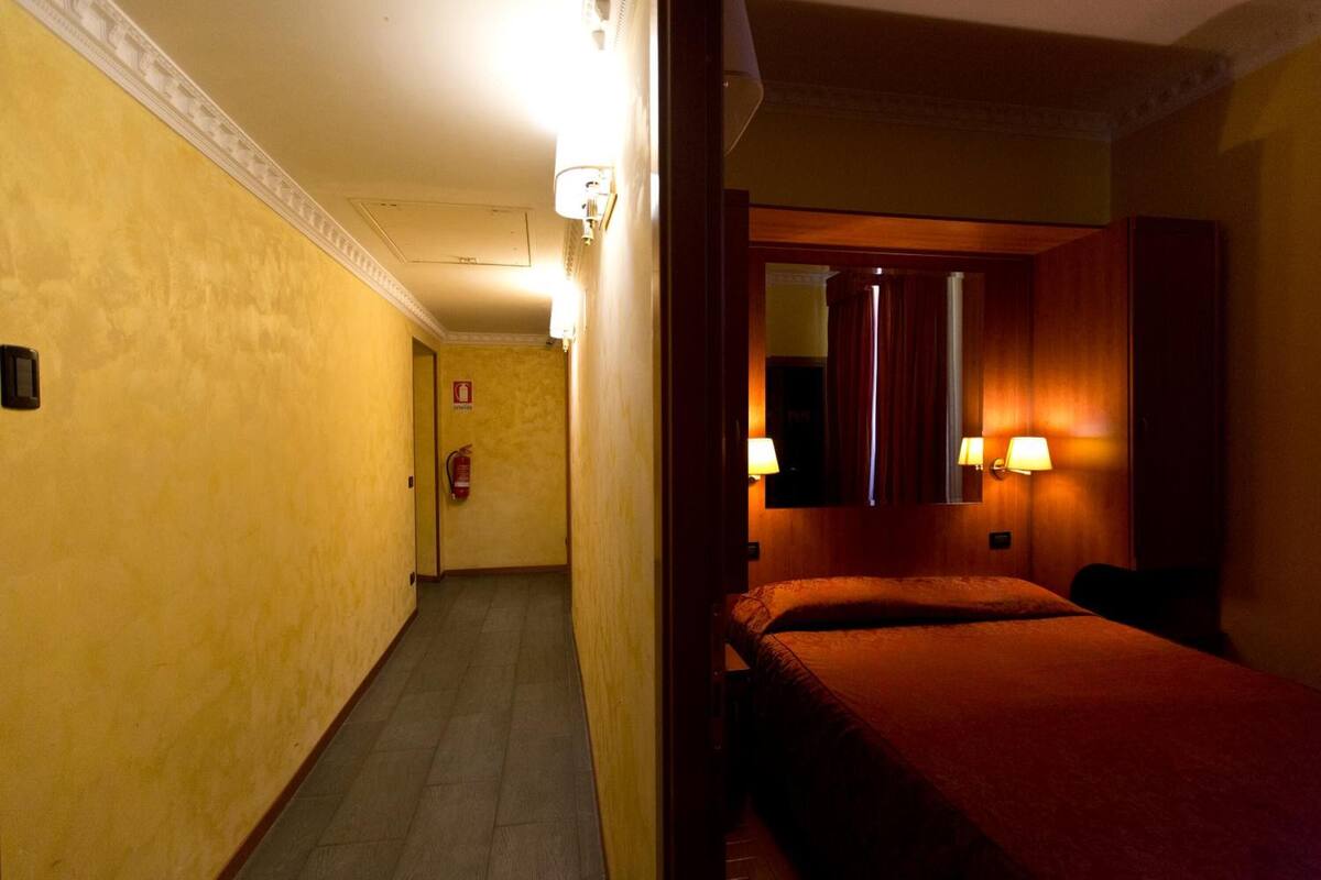 Quarto de hotel com cama, mesa de cabeceira, espelho e luminária na parede.