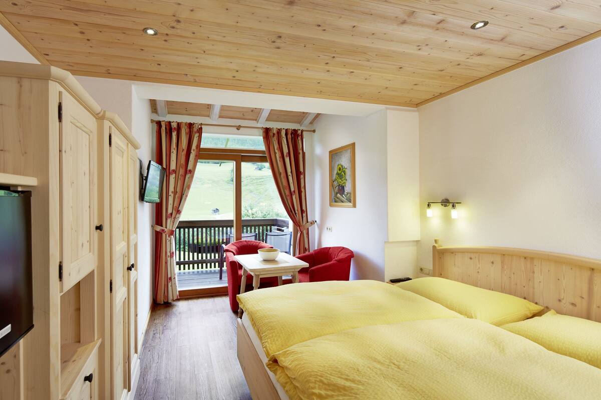 A room with two beds, a TV, wooden cabinets, a balcony with red curtains, and a painting on the wall.