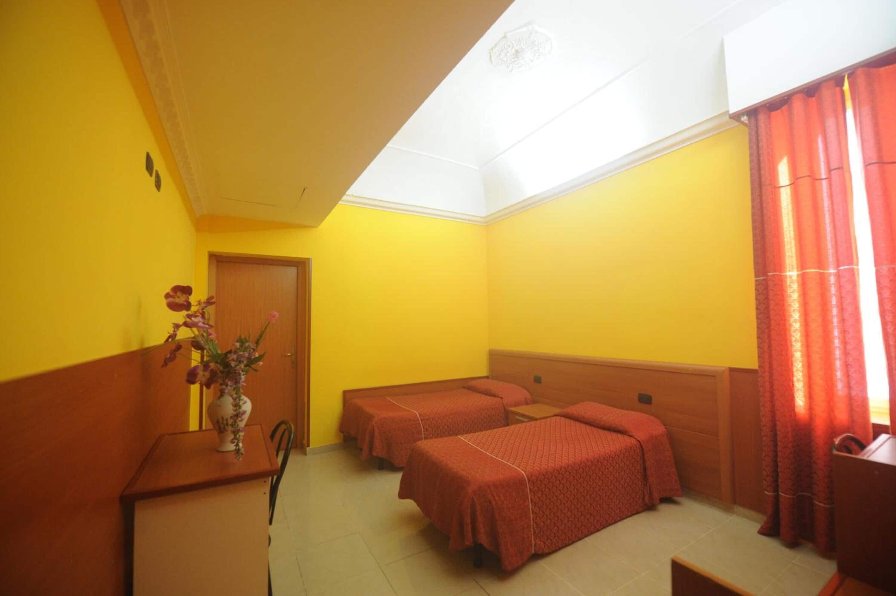 A room with two beds, a wooden desk with a vase of flowers, and a door.
