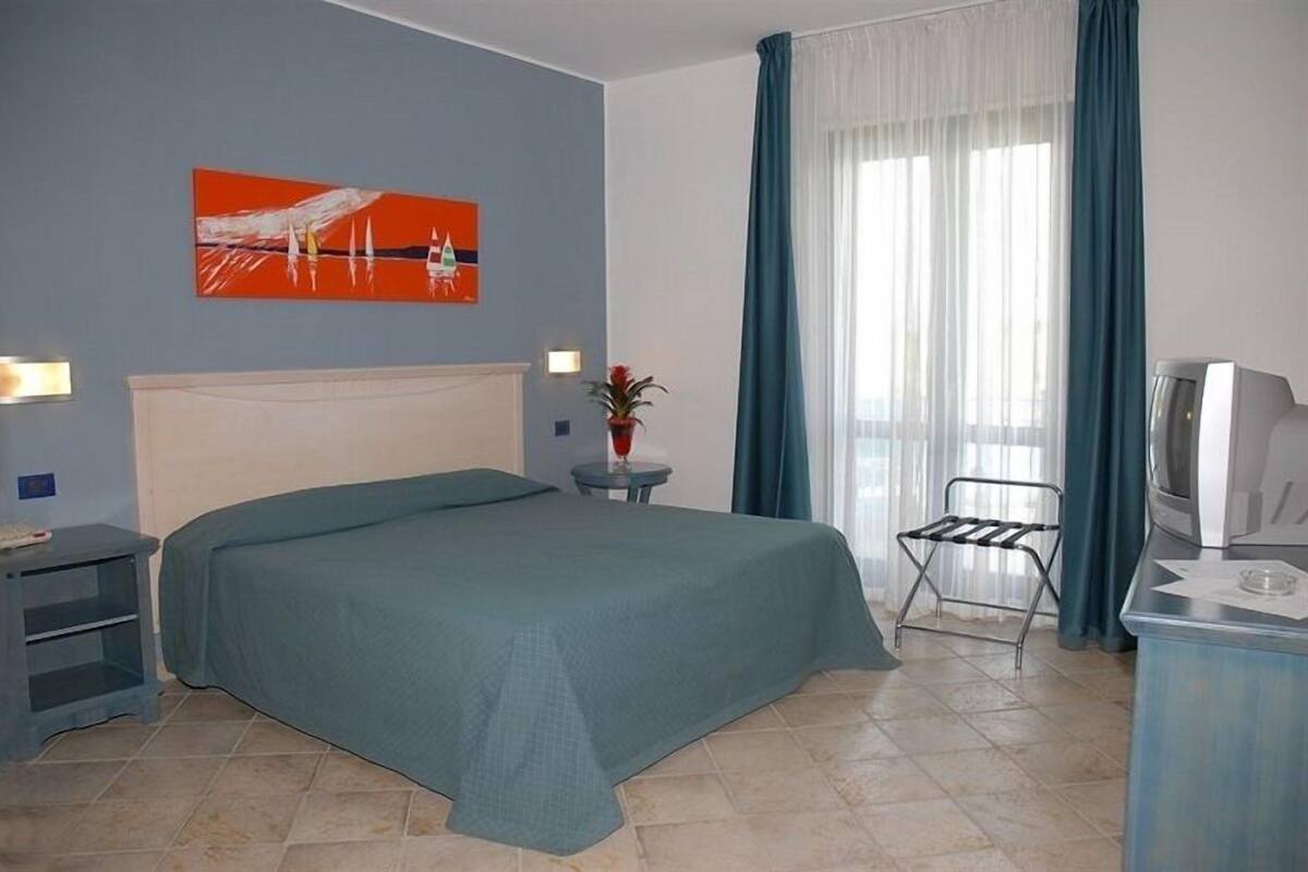 A hotel room with a bed, a small table with a plant, a chair, a desk with a computer, and a window with curtains.