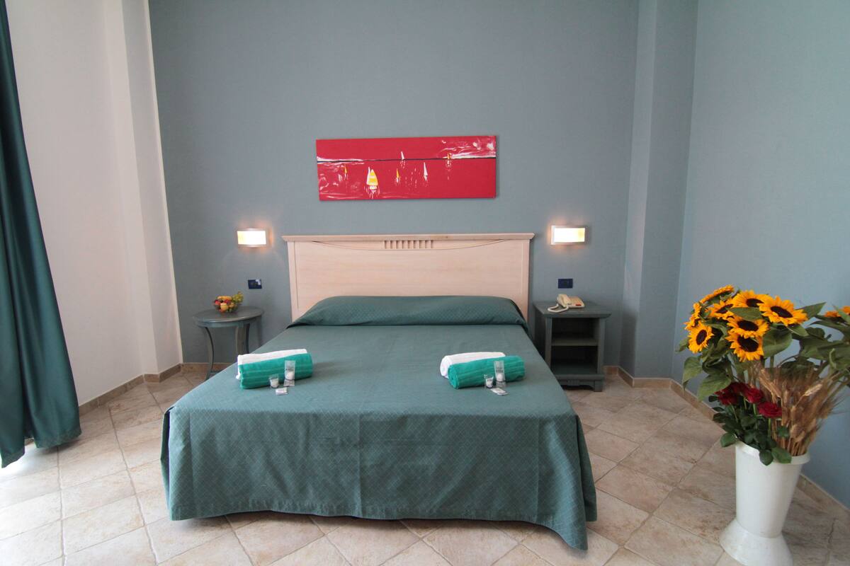 A neatly made bed with a teal bedspread, two pillows, and two folded towels. There is a red abstract painting on the wall above the bed, and a vase with sunflowers on the floor to the right.