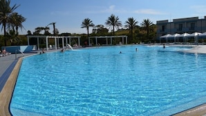 Une piscine d’un bleu éclatant, bordée de chaises longues et d’ombrelles, entourée de palmiers et d’un bâtiment doté de nombreuses fenêtres.