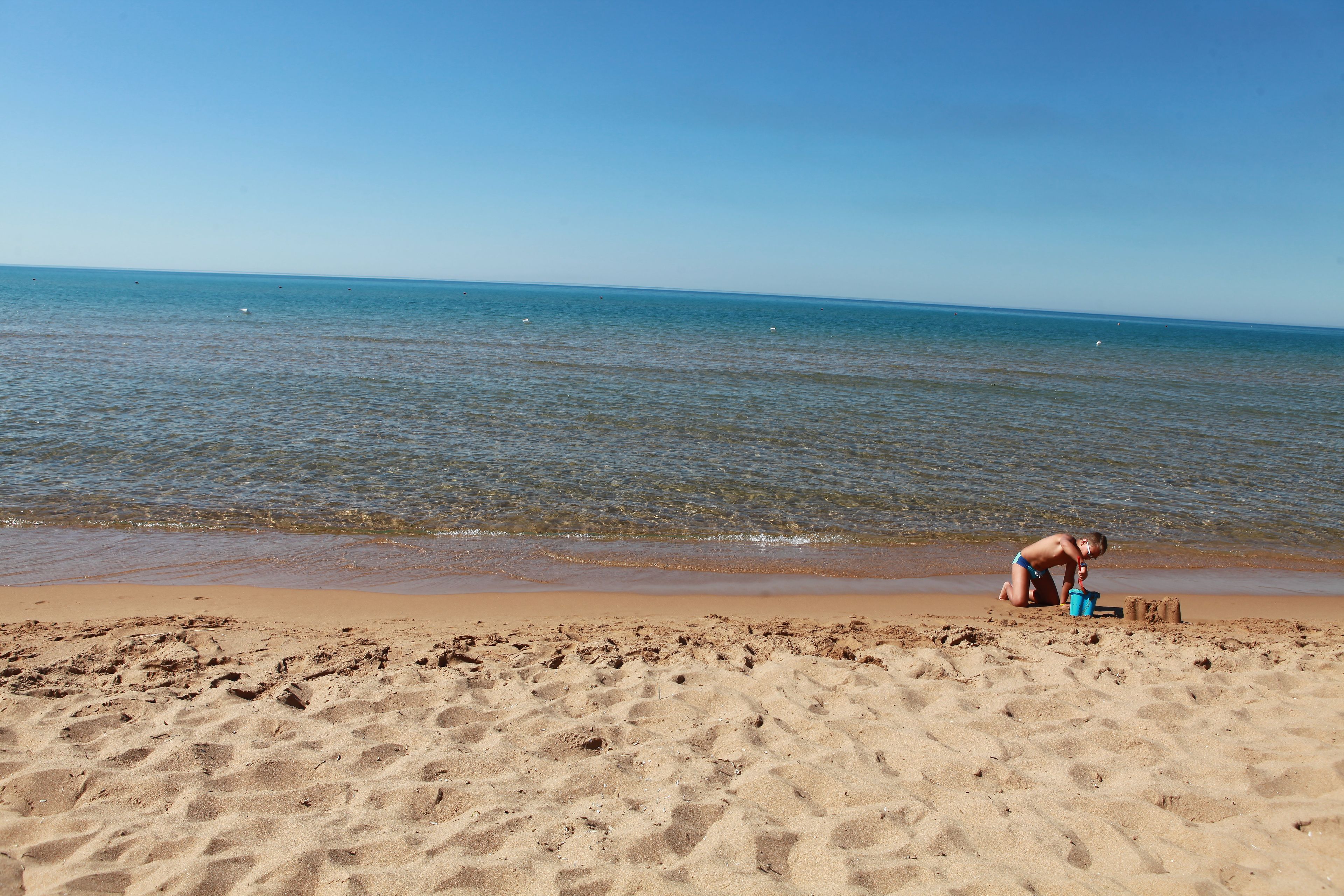 A sandy beach with footprints leading to the water, a child playing with a blue bucket, and a clear blue sky.