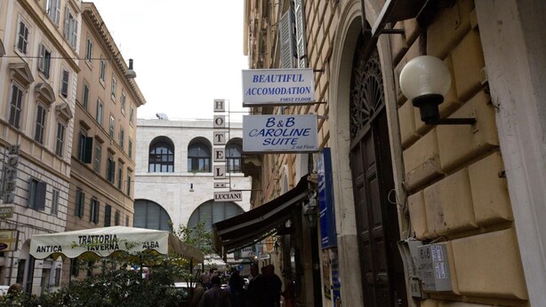 A narrow street with historic buildings, signs for 'Beautiful Accommodation' and 'B&B Caroline Suite' on the first floor, and 'Trattoria Taverna' on the ground floor. - Hostel Beautiful (Rome)