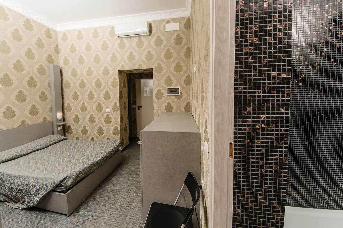 A compact room with a bed, a small desk, and a chair. There is a door leading to another room and a window with a mesh screen.