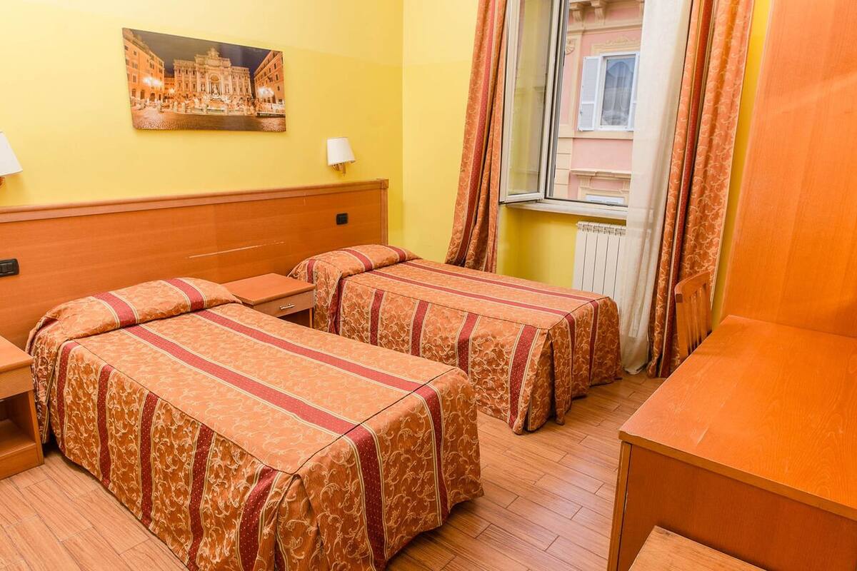 A hotel room with two beds, a wooden headboard, a table, a window with curtains, and a painting on the wall.