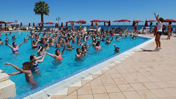 Zona de piscina con varias personas nadando, tumbonas y sombrillas rojas.