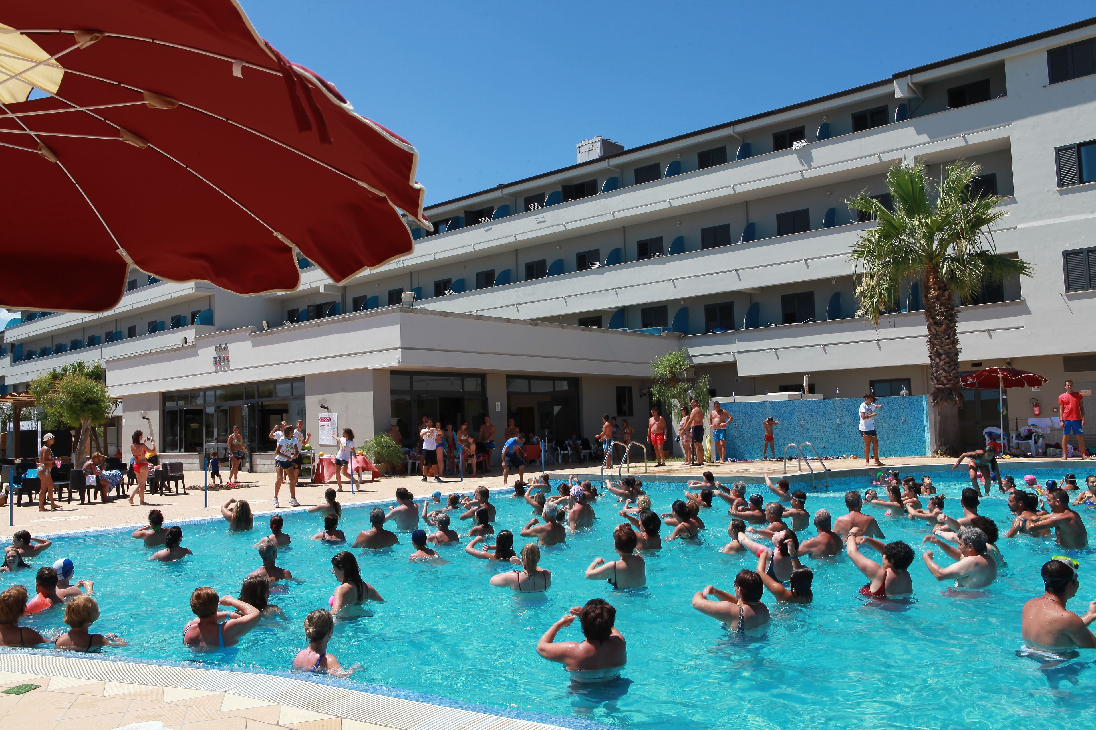 Una piscina con persone che si godono l'acqua, circondata da un edificio con numerosi balconi e una palma.