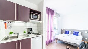 Una camera d'albergo compatta con angolo cottura, un letto con cuscini blu e una finestra con tende viola.