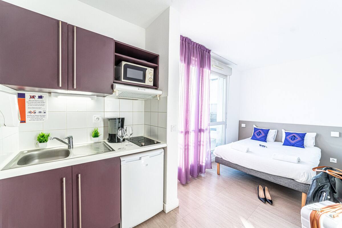 Ein kompaktes Hotelzimmer mit einer Küchenzeile, einem Bett mit blauen Kissen und einem Fenster mit violetten Vorhängen.